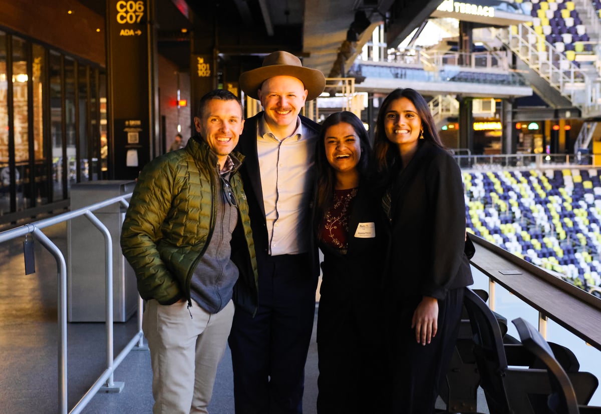 Four colleagues smiling for a photo in a stadium suite overlooking the seating area.