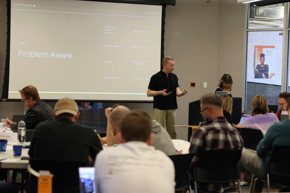 A presenter stands before a large projector screen showing a marketing slide while an audience listens in a meeting room