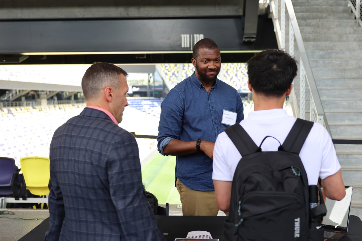 Three men talking at a table in a stadium concourse, with the man in the center facing the camera.