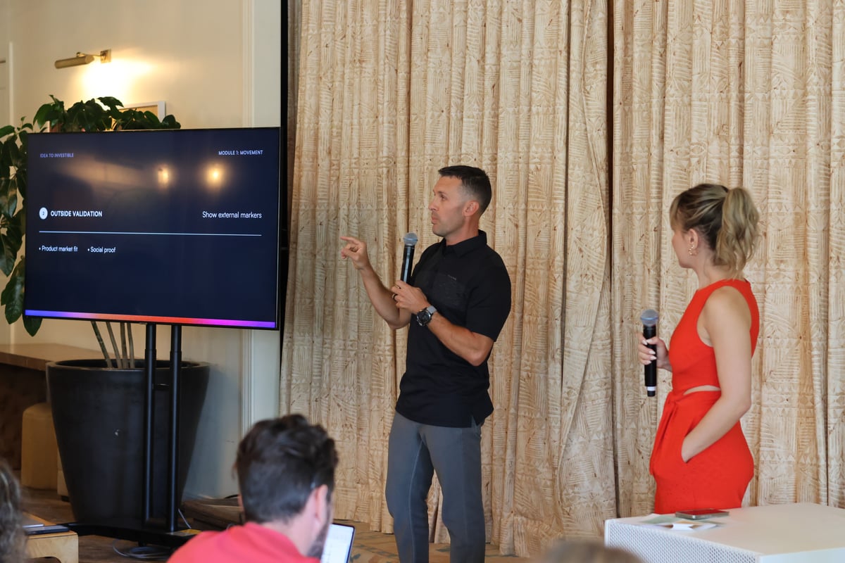 A man in a black polo shirt giving a presentation to an audience while a woman in a red dress stands beside him.