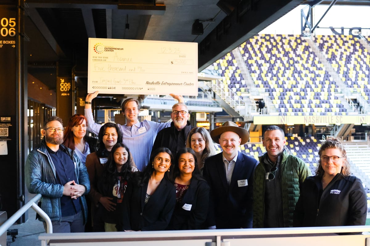 A diverse group of ten people stands in a stadium concourse holding a large ceremonial check for $5,000.