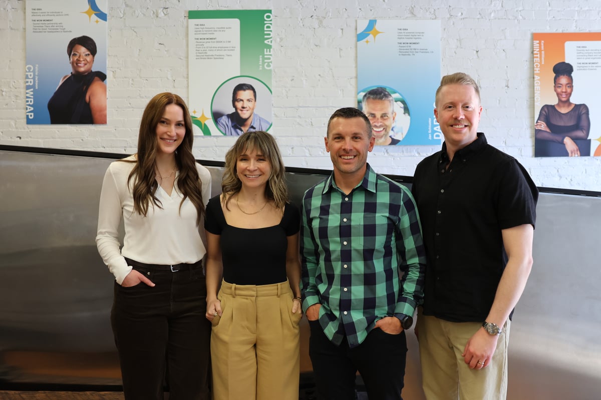Four people standing in a row in front of a white brick wall decorated with posters of entrepreneurs.